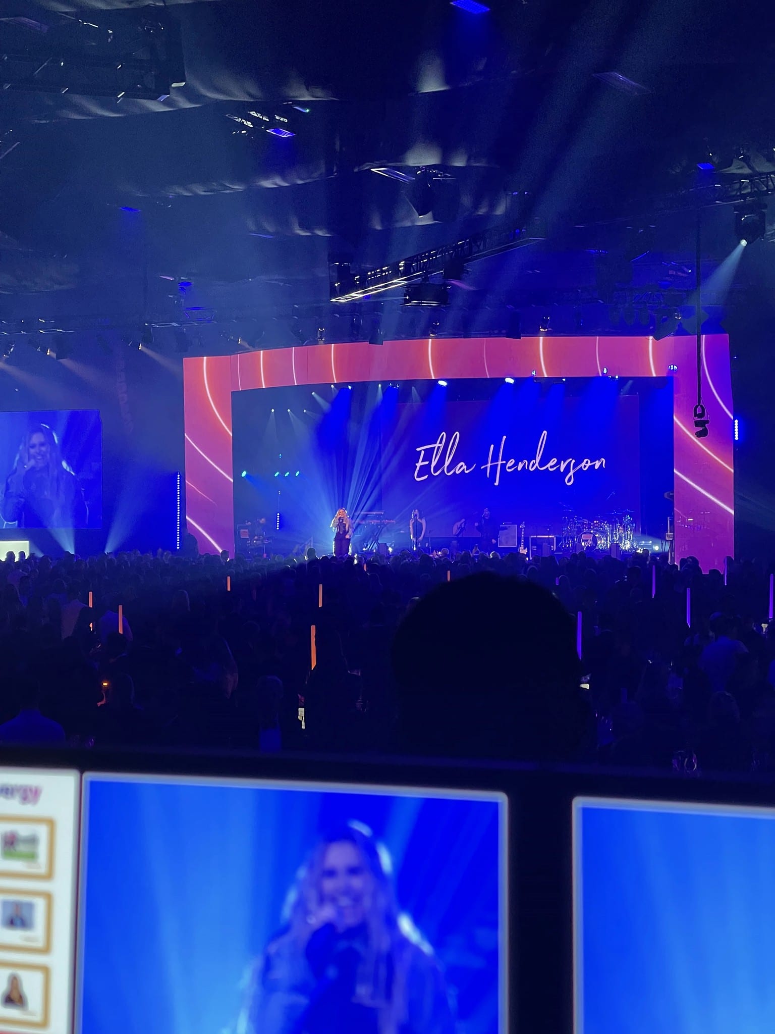 Ella Henderson on stage, IMAG and media-server monitors in the foreground, Sainsbury's Big Bash, Evolution London, December 2025.
